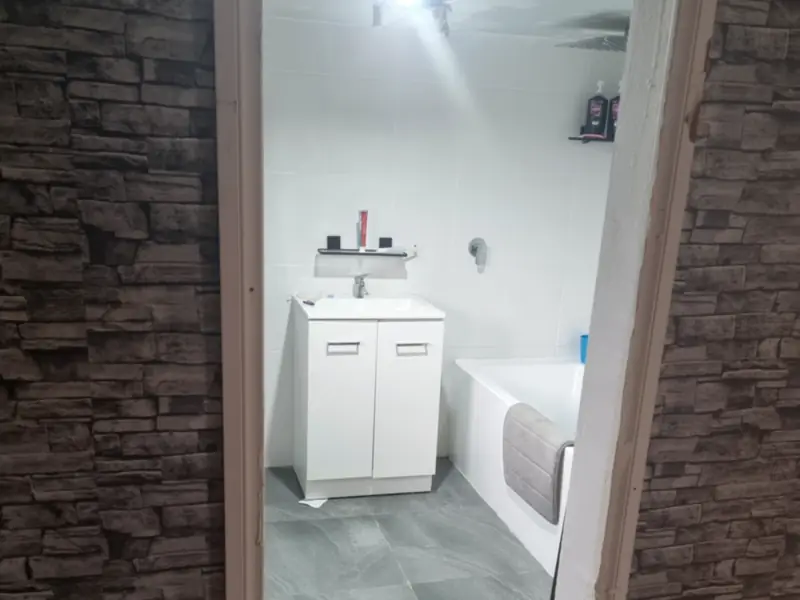 A white bathroom with a vanity, bath and grey tiled floor, seen through a doorway in a dark stacked-stone wall