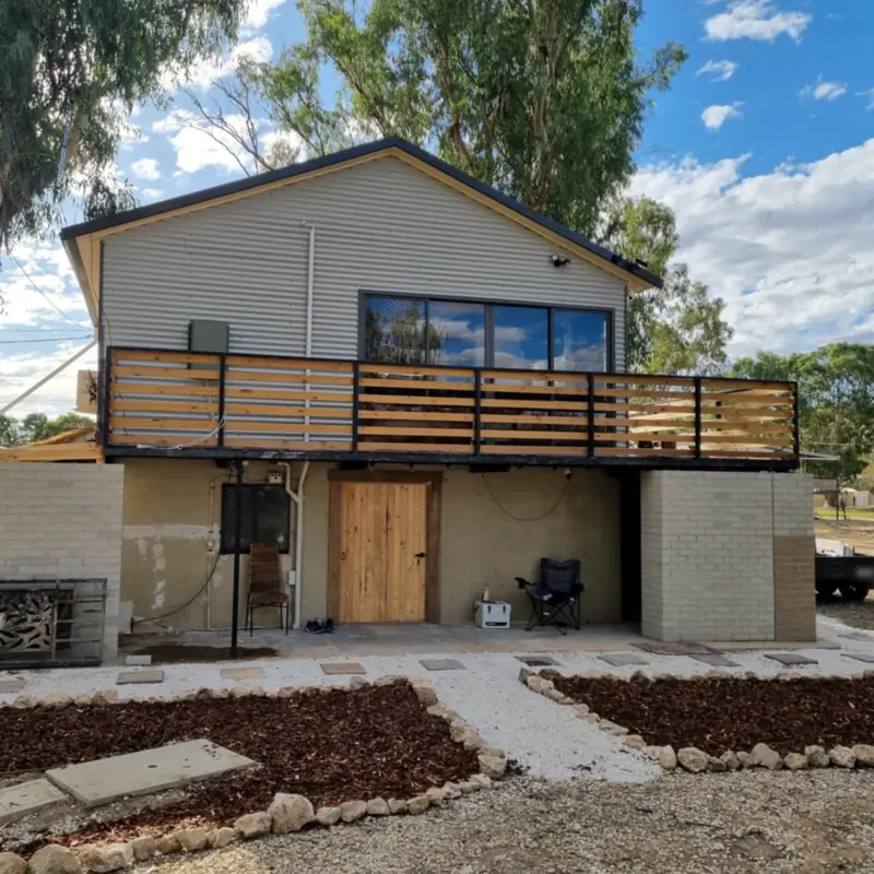A raised house with corrugated iron cladding, a timber-slat deck, a painted brick base, gravel paths and garden beds