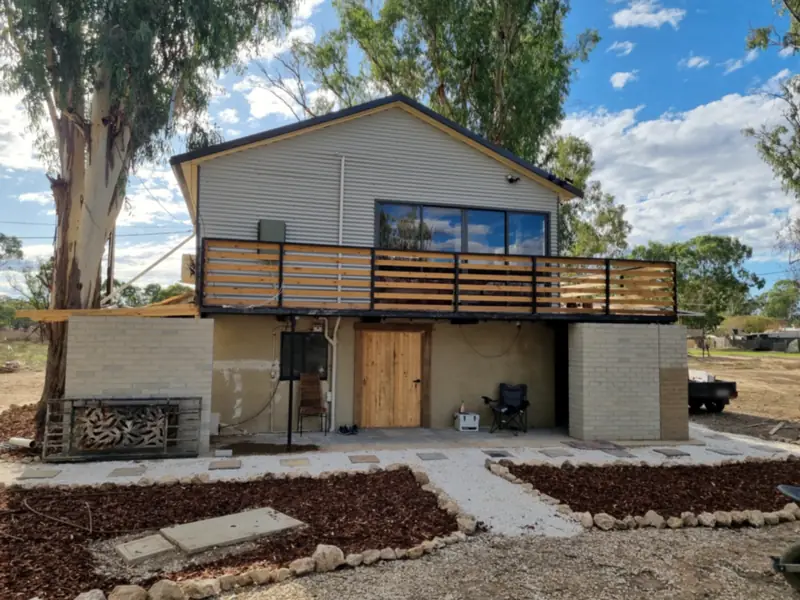A raised house with corrugated iron cladding, a timber-slat balcony, a painted brick base and new garden beds edged in stone