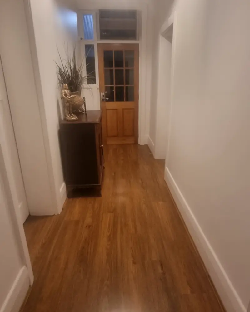 A finished hallway: fresh white walls and skirting boards, a polished timber floor, and an old glazed door and transom at the end