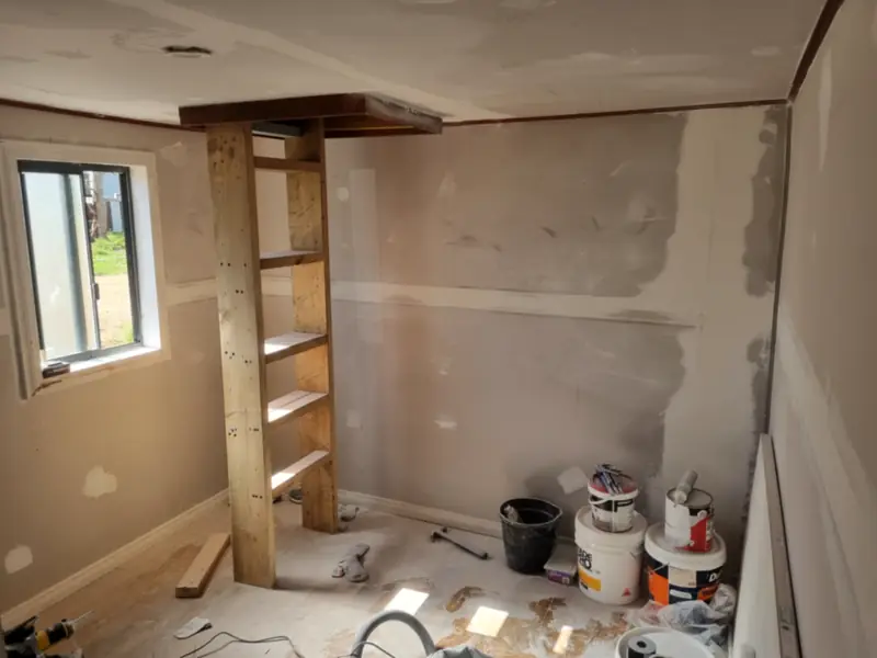 A room with plaster walls set and ready to sand, a timber shelf unit and paint tins