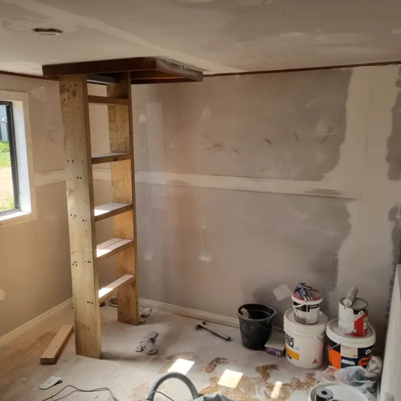 A room part-way through a job: plaster walls set and ready to sand, a timber shelf unit, and paint tins on the floor